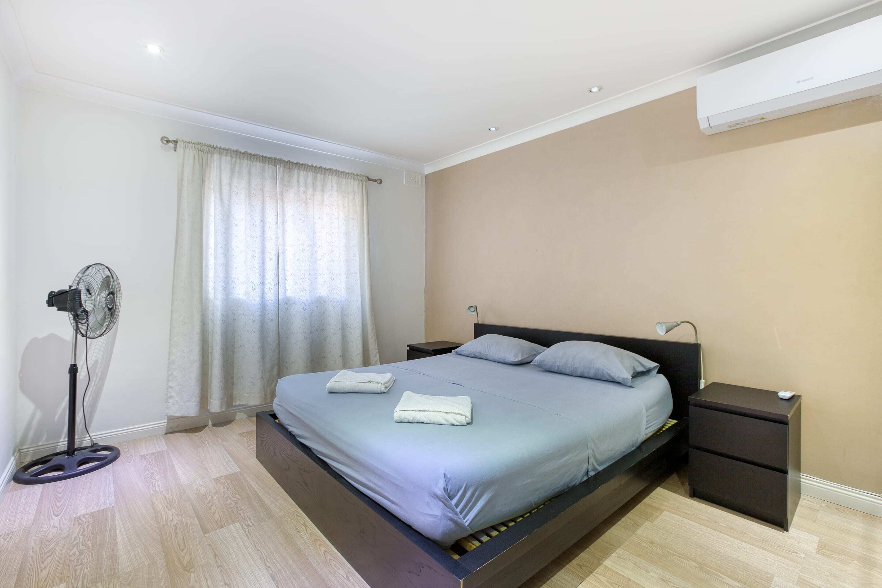 A bedroom with a bed, bedside tables, a standing fan, and a window with curtains.