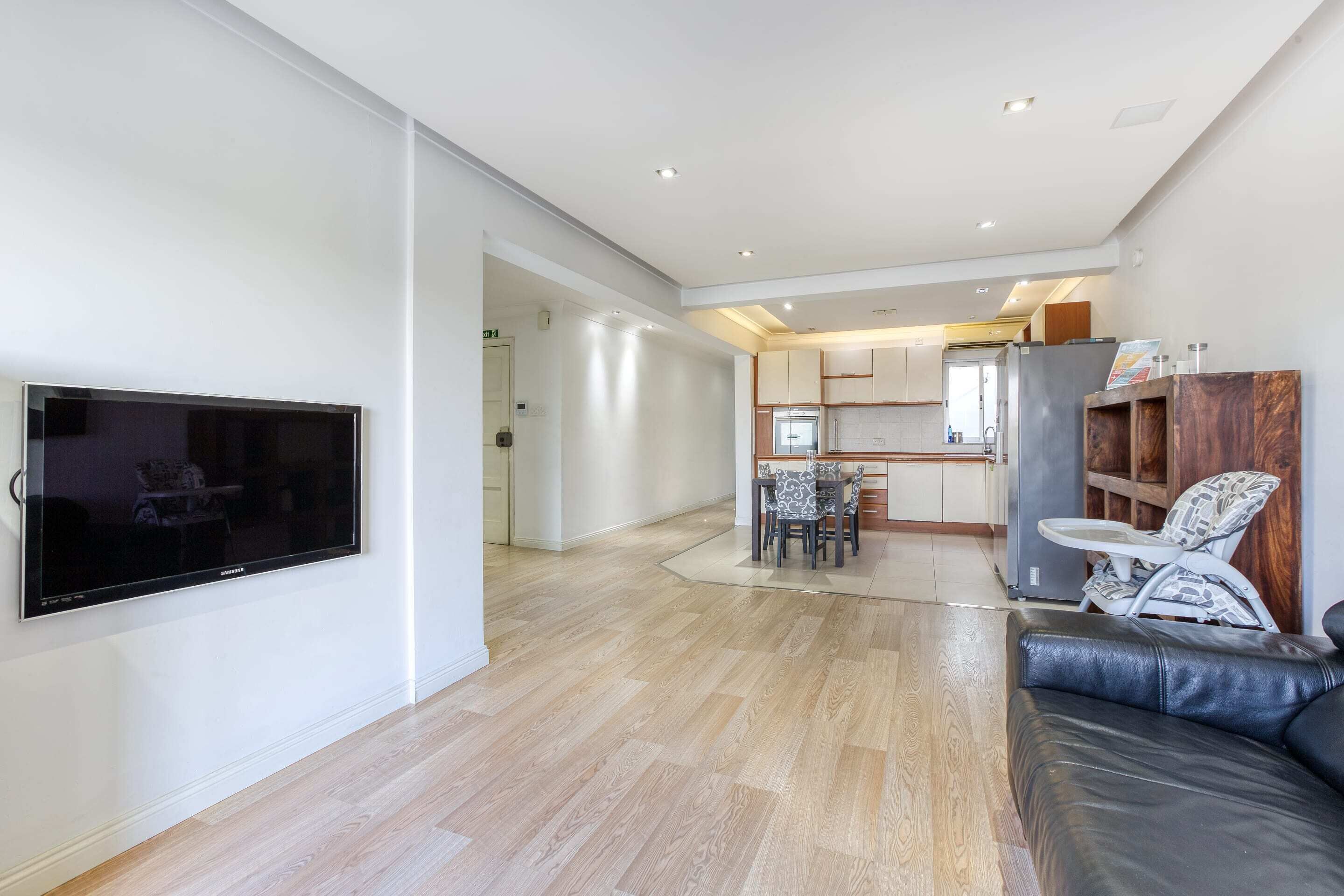 A modern living room with a flat-screen TV, a leather sofa, a dining area with a table and chairs, and a kitchen in the background.