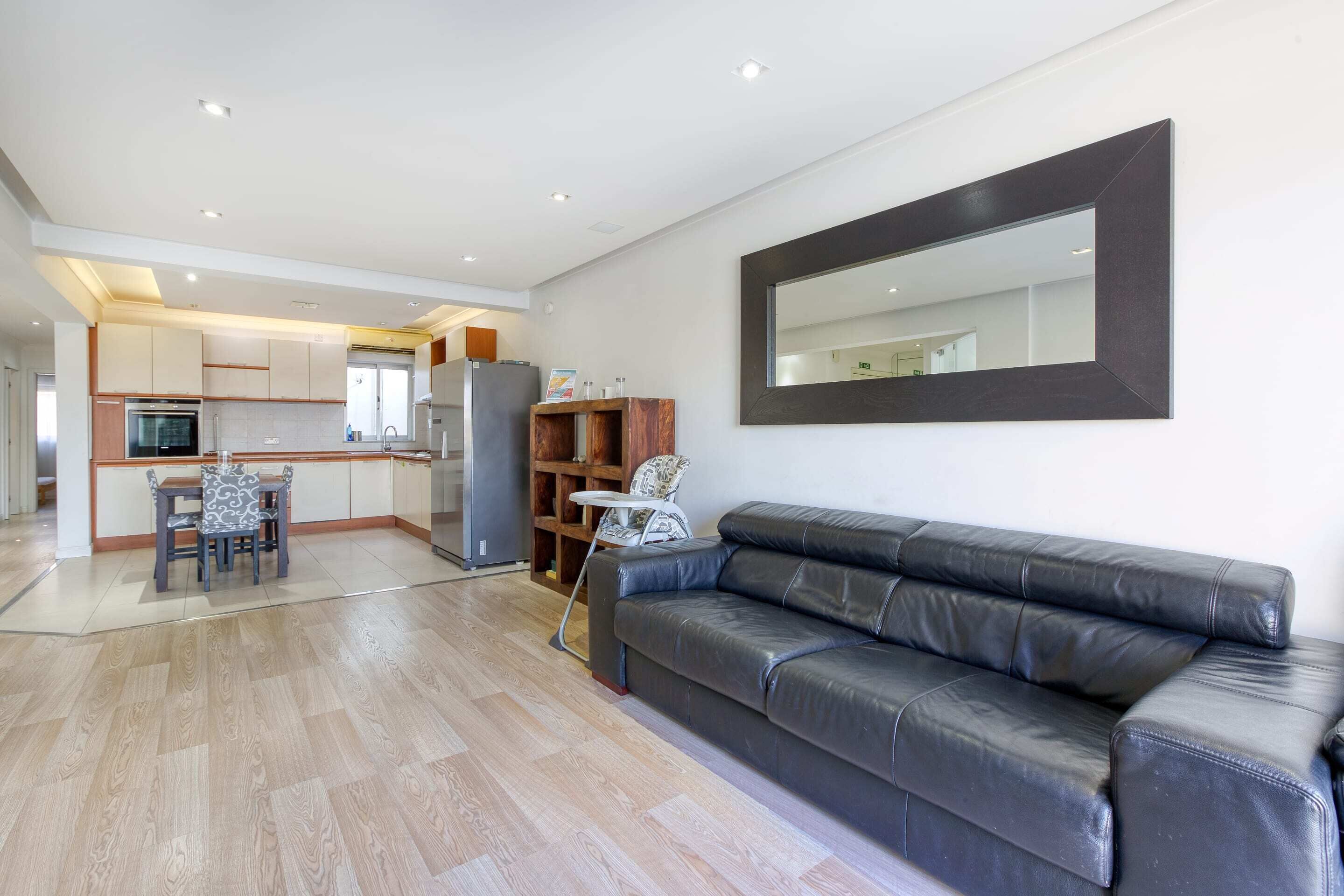 Un salon moderne avec un canapé en cuir noir, une table à manger avec des chaises, et un coin cuisine à l’arrière-plan.