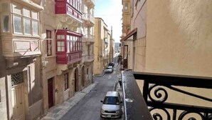 A narrow street flanked by multi-story buildings with balconies, a blue car parked on the side, and a clear sky. - Valletta Studio (Valletta)