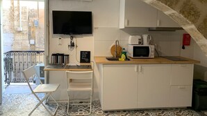 A compact kitchen with a microwave, kettle, and a small sink. There's a TV mounted on the wall, a chair, and a welcome sign. The room has a tiled floor and a stone archway. - Valletta Studio (Valletta)
