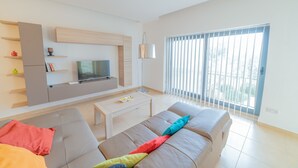 A modern living room with a flat-screen TV, a sofa, a coffee table, and a large window with blinds. - Luxury Seaview & Pool (Mellieha)