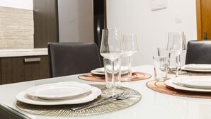Mesa de comedor dispuesta con platos blancos, cubiertos y copas de vino, con un interior moderno y vistas al exterior.