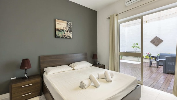 Habitación de hotel moderna con una cama grande, una mesita de noche, una lámpara y vistas al balcón exterior.