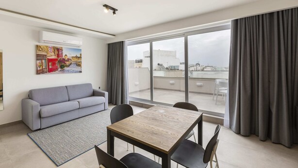 A modern living room with a grey sofa, a wooden dining table with four chairs, a large window with grey curtains, and a cityscape view. - Gzira Suite 13 (Gzira)