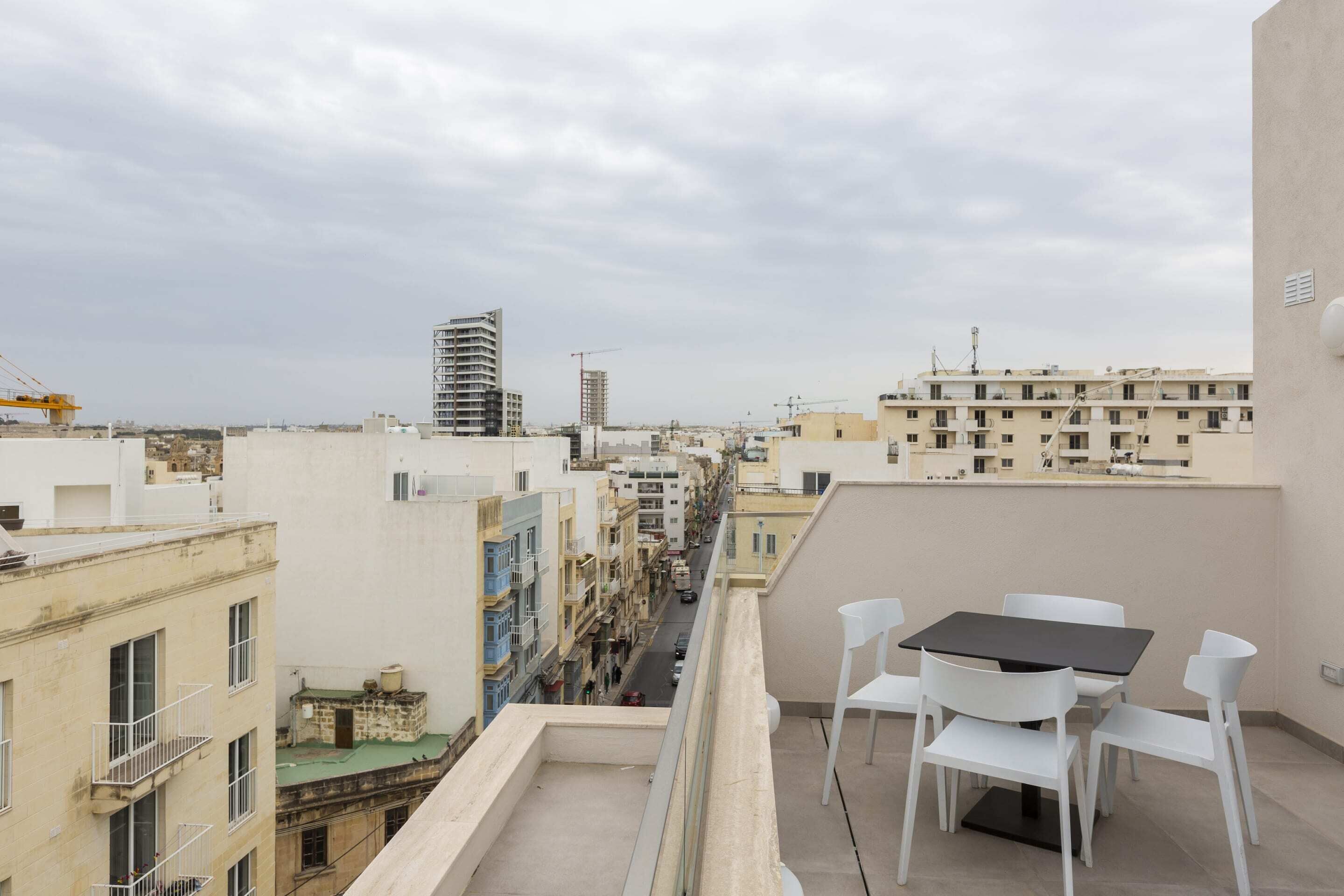 Un balcon avec une table et des chaises, donnant sur une rue bordée d’immeubles et où l’on aperçoit, au loin, une grue de chantier.