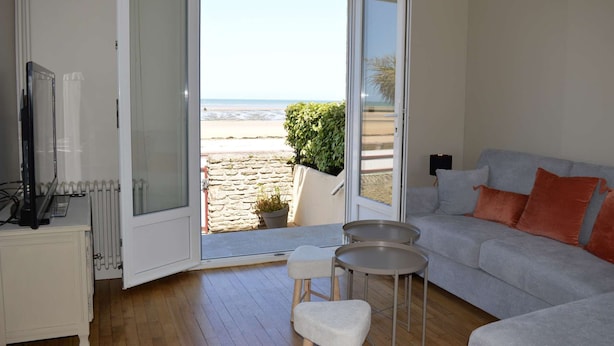 A living room with a sofa, two small stools, a TV, and a view of the beach through a glass door. - L'estran - Maison sur la Digue Avec vue mer (Courseulles-sur-Mer)
