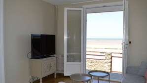 A room with a television on a cabinet, a small table, a sofa, and a sliding glass door leading to a beach view. - L'estran - Maison sur la Digue Avec vue mer (Courseulles-sur-Mer)