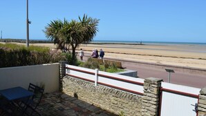 A stone wall with a metal gate, a small table and chairs, and a view of a beach with people walking. - L'estran - Maison sur la Digue Avec vue mer (Courseulles-sur-Mer)