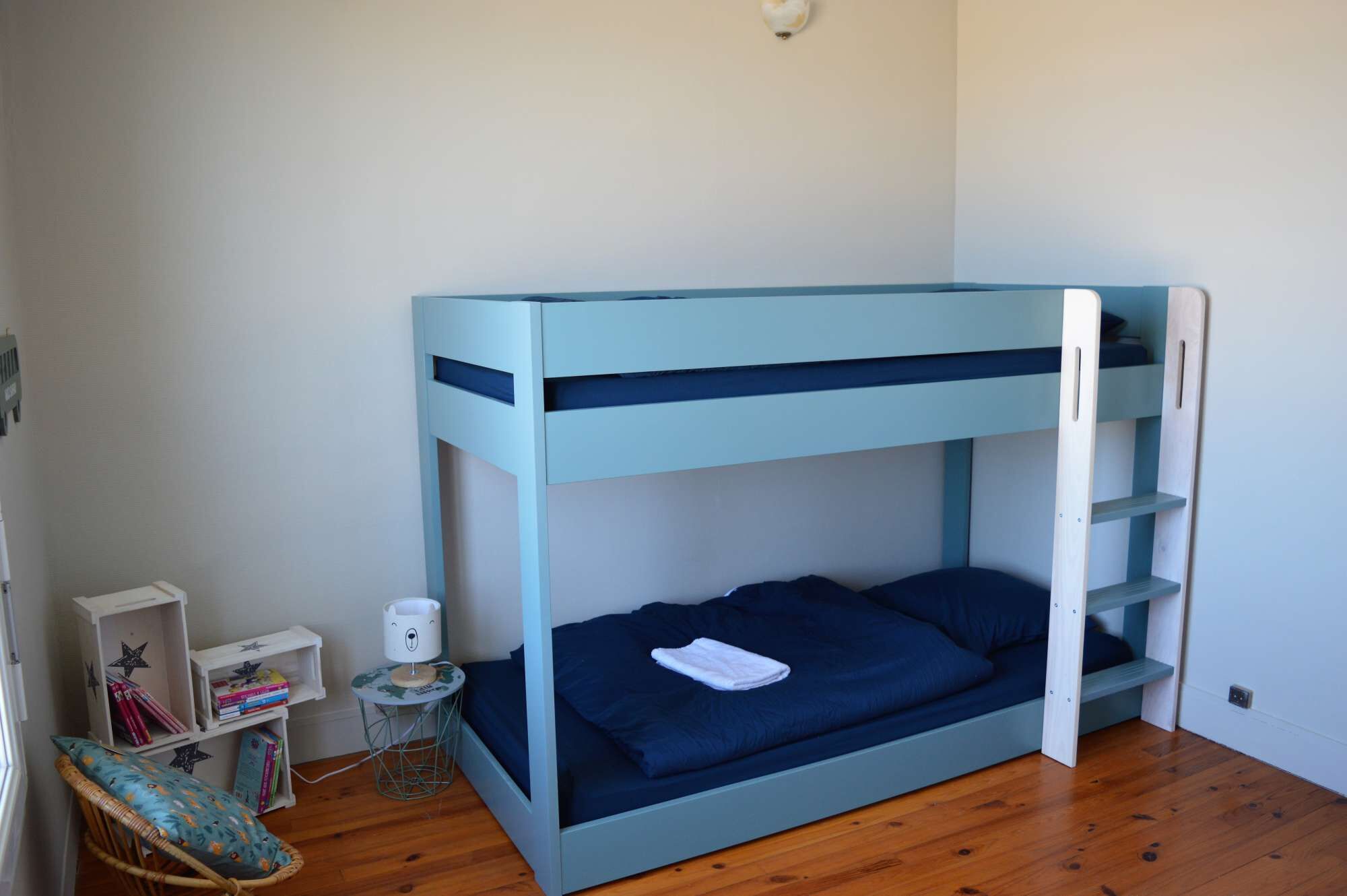 Ein Stockbett mit blauer Rahmen und einem Bett darunter. Ein kleiner Tisch mit Lampe, Stuhl und Bücherregal mit Büchern.