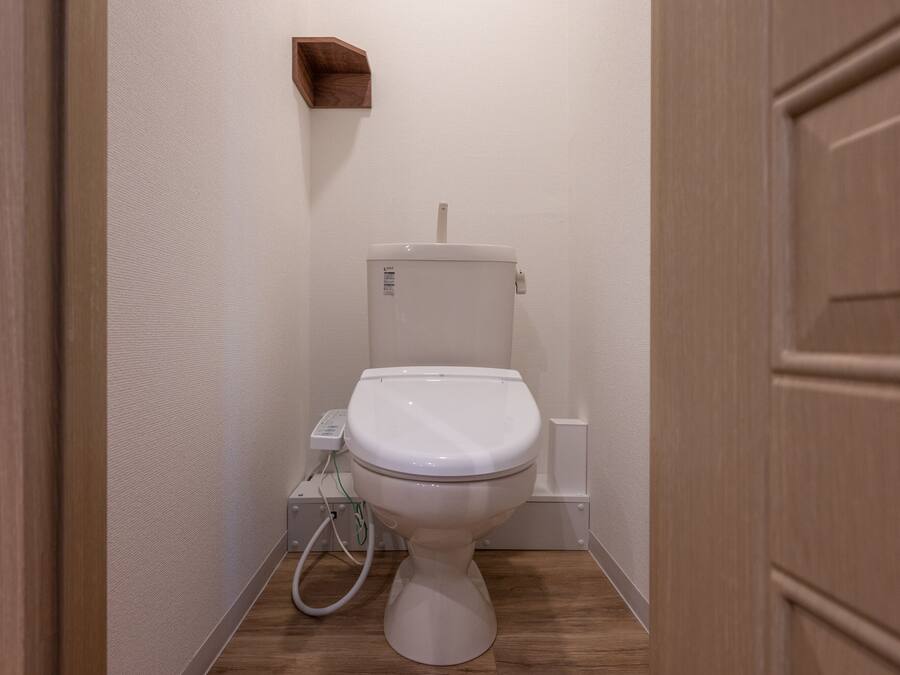 Un baño pequeño y limpio, con inodoro blanco, piso de madera y estante fijado a la pared.