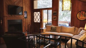 A cozy cabin interior with a fireplace, a sofa, a television, and a large window. - Escape to Ptarmigan Village 64 (Whitefish)