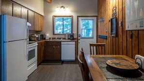 A compact kitchen with wooden cabinets, a refrigerator, a microwave, a sink, and a dining area with a wooden table and chairs. - Escape to Ptarmigan Village 64 (Whitefish)