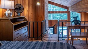 Interno di una baita in legno con un letto, una cassettiera, una ventola, una lampada e una statuetta di un orso.