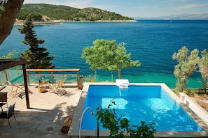 A pool with a view of the sea, surrounded by trees and a wooden deck with chairs and a table.