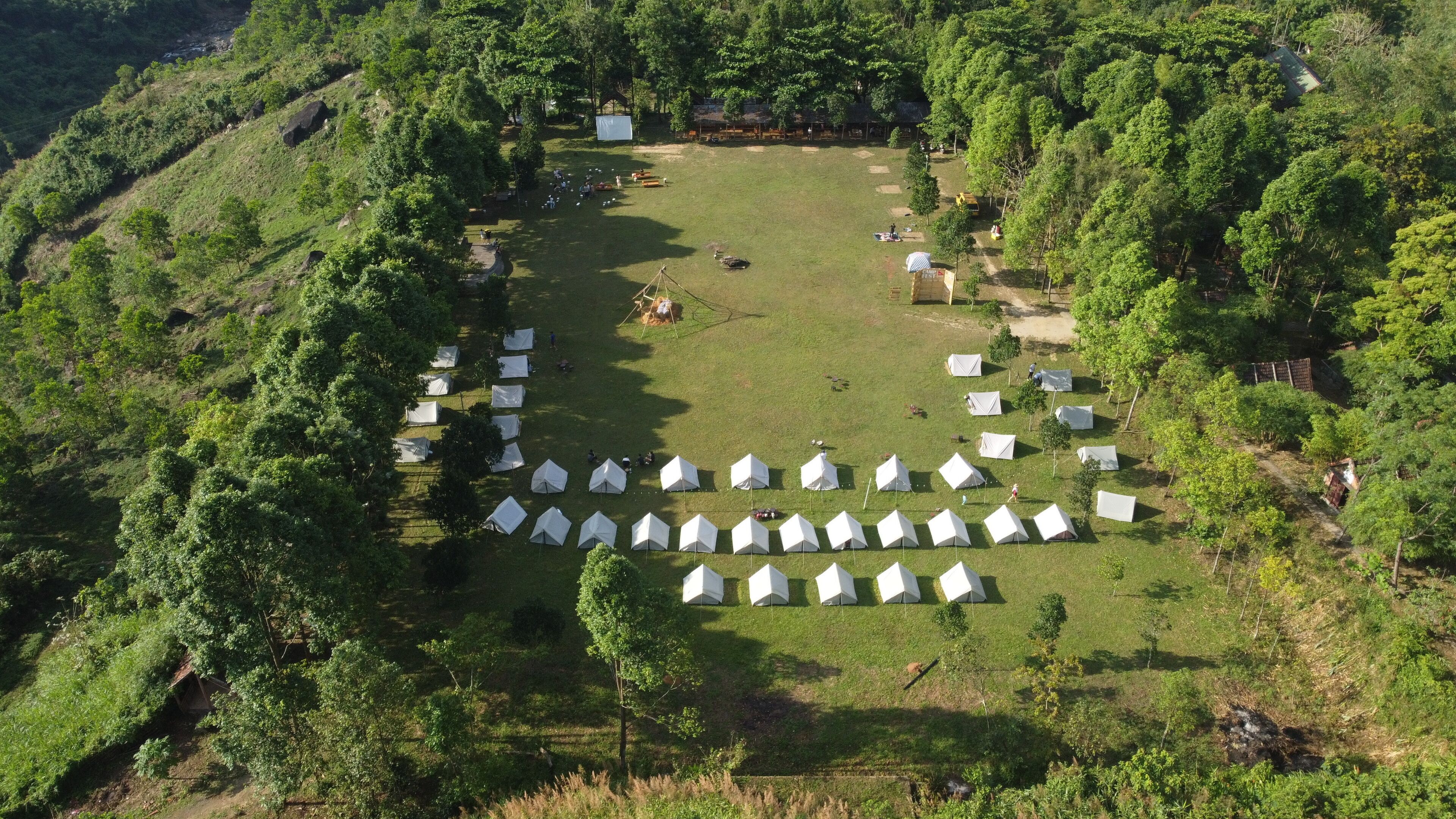 Vista aérea de un camping con varias tiendas de campaña dispuestas en círculo sobre un campo de césped rodeado de árboles frondosos.