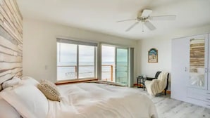 A bedroom with a bed, a ceiling fan, a wardrobe, and a view of the sea through large windows.