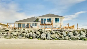 A two-story beach house with a balcony, large windows, and a stone retaining wall.