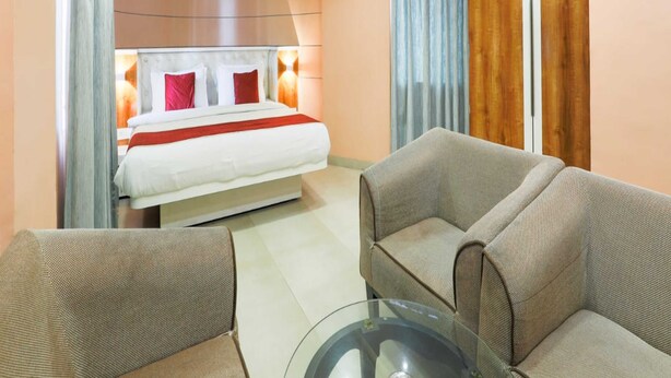 Ein Hotelzimmer mit einem Bett, zwei Sesseln, einem Glastisch, einem großen Fenster mit Vorhängen und einem Deckenventilator.