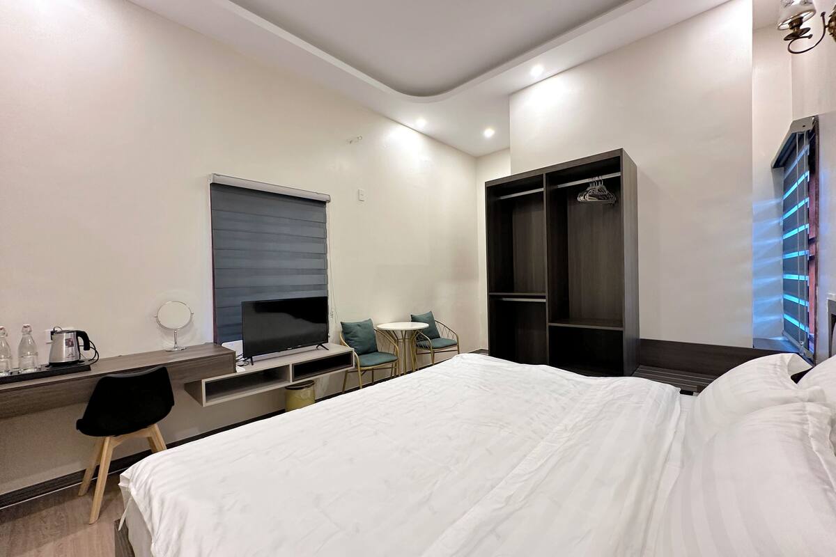 A modern hotel room with a large bed, a desk with a flat-screen TV, a seating area with chairs, and a wardrobe with a mirror.