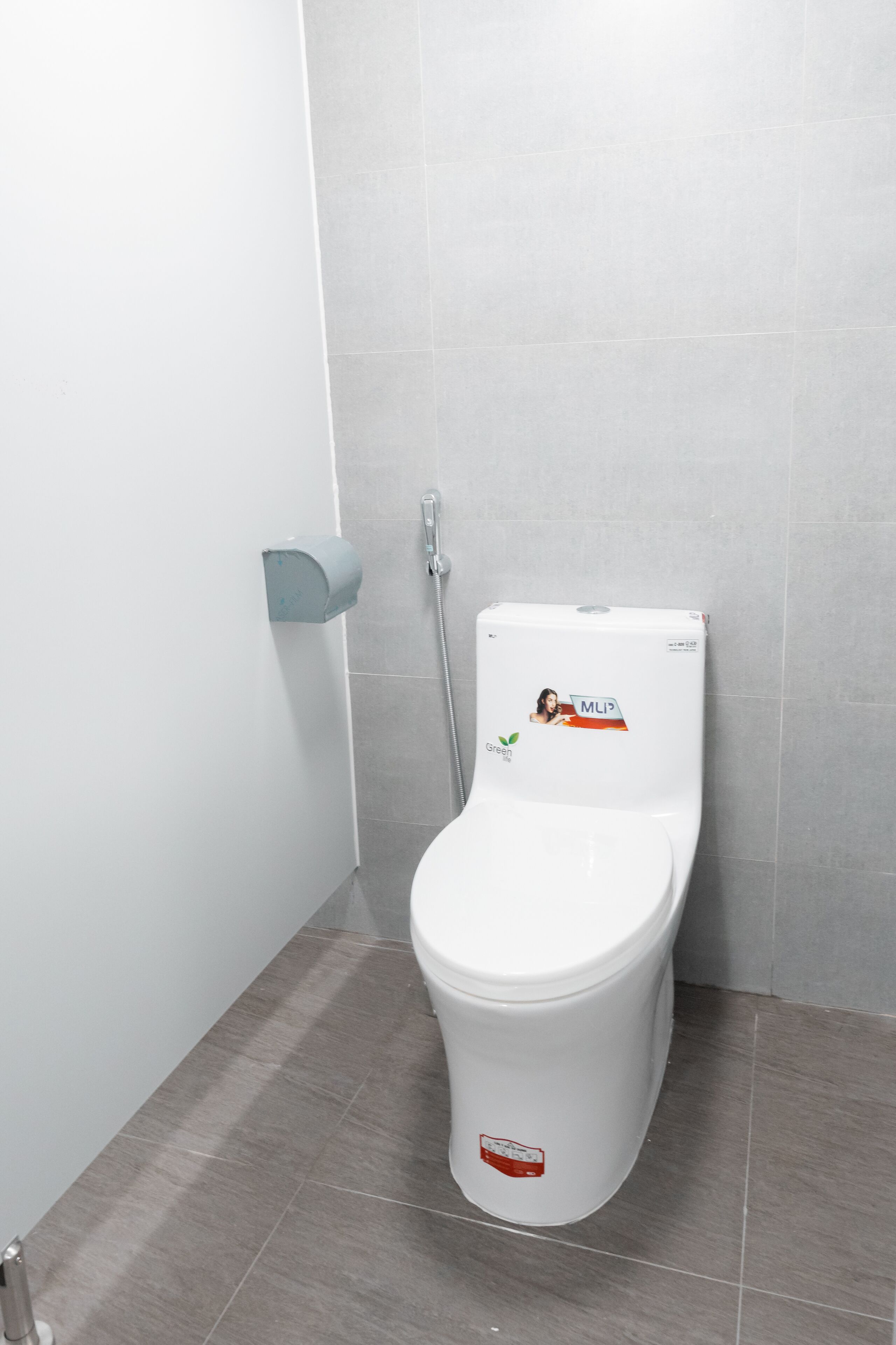 Inodoro blanco con una pegatina verde, dispensador de jabón empotrado en la pared y suelo con baldosas grises.
