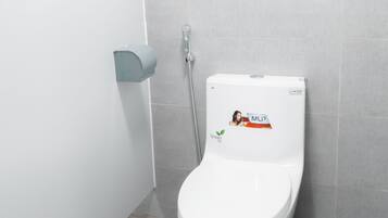 Inodoro blanco con una pegatina verde, dispensador de jabón empotrado en la pared y suelo con baldosas grises.