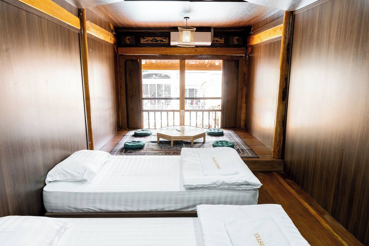 A room with two beds, a small wooden table, and a view of a balcony with a railing.