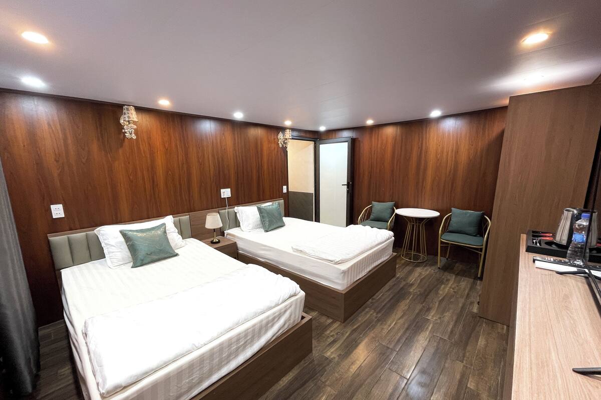 A hotel room with two beds, a small round table, a chair, and a desk with a kettle and remote control.