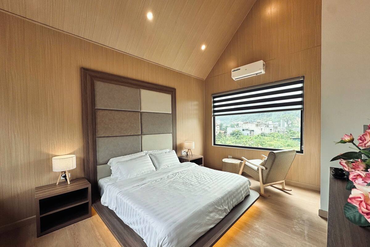 A modern bedroom with a large bed, a bedside table, a chair, a window with blinds, and a view of greenery outside.
