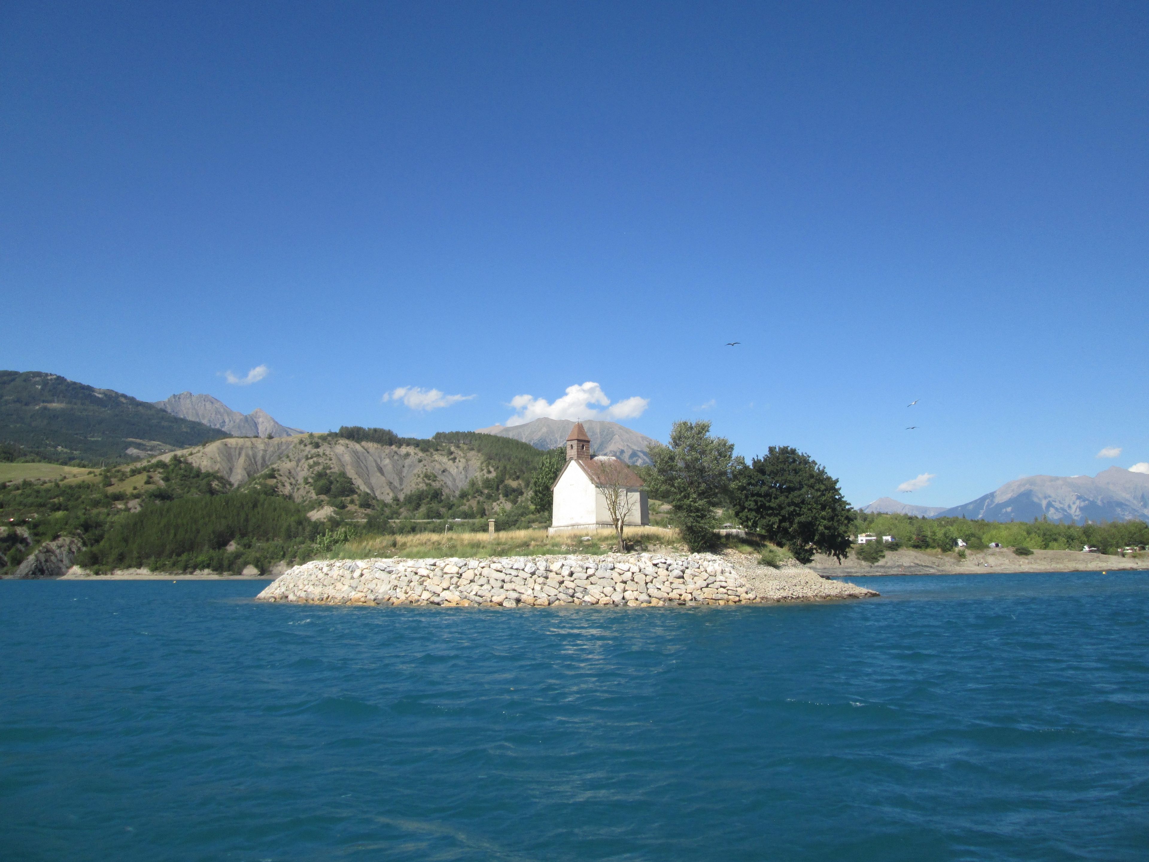 A small white building on a rocky island surrounded by water, with mountains in the background.