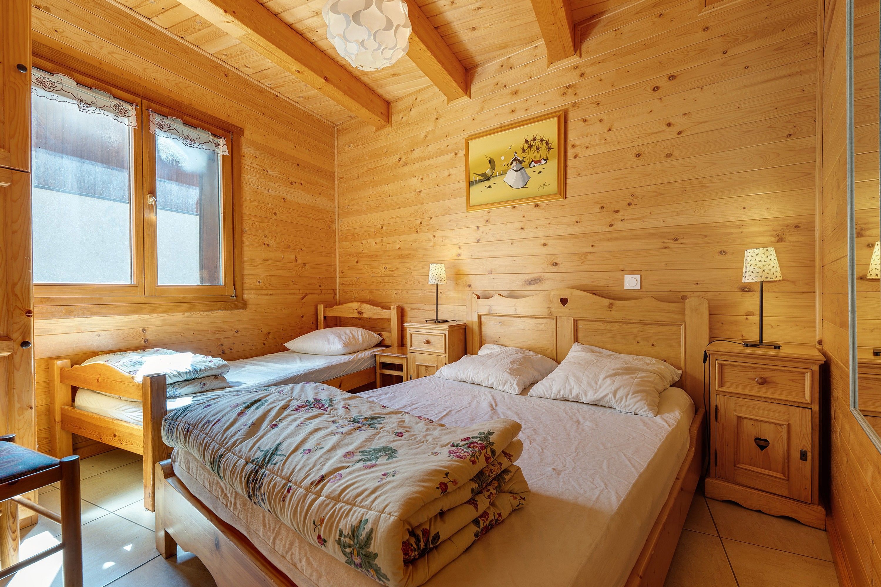 Une chambre dans un chalet en bois, avec deux lits, une fenêtre, un tableau et une table de chevet avec une lampe.