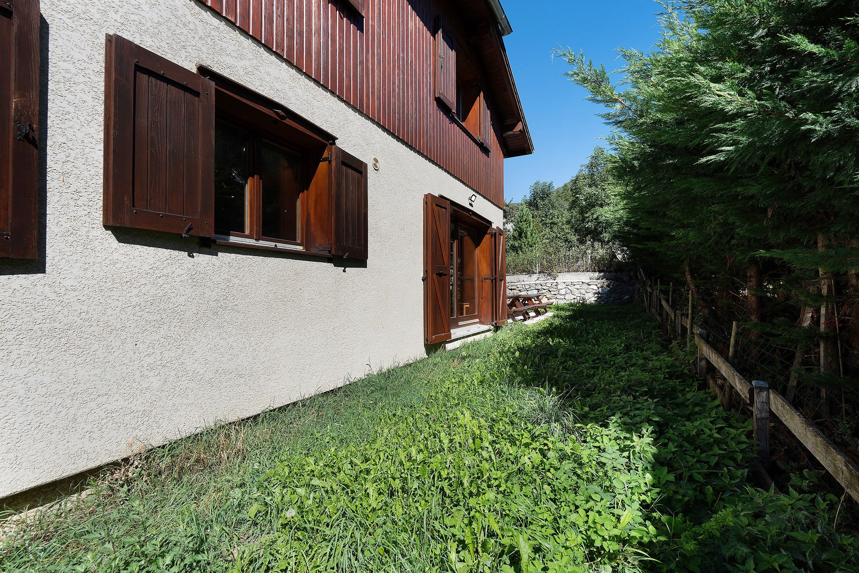Un bâtiment à deux étages, doté de volets en bois, d’un mur en pierre et d’un espace verdoyant avec une végétation dense.