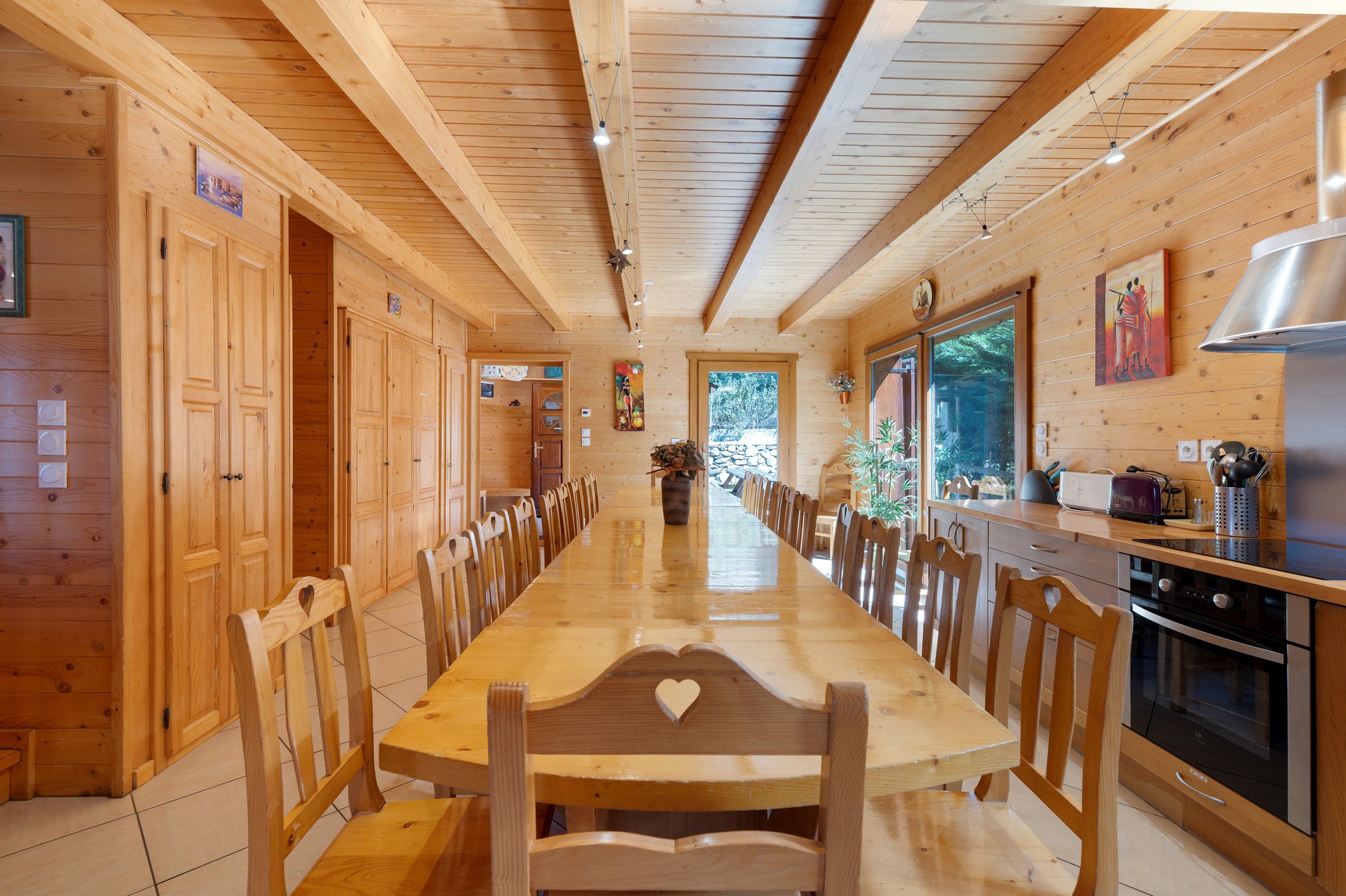 Interior em madeira com uma longa mesa de jantar, cadeiras, área de cozinha e uma grande janela.