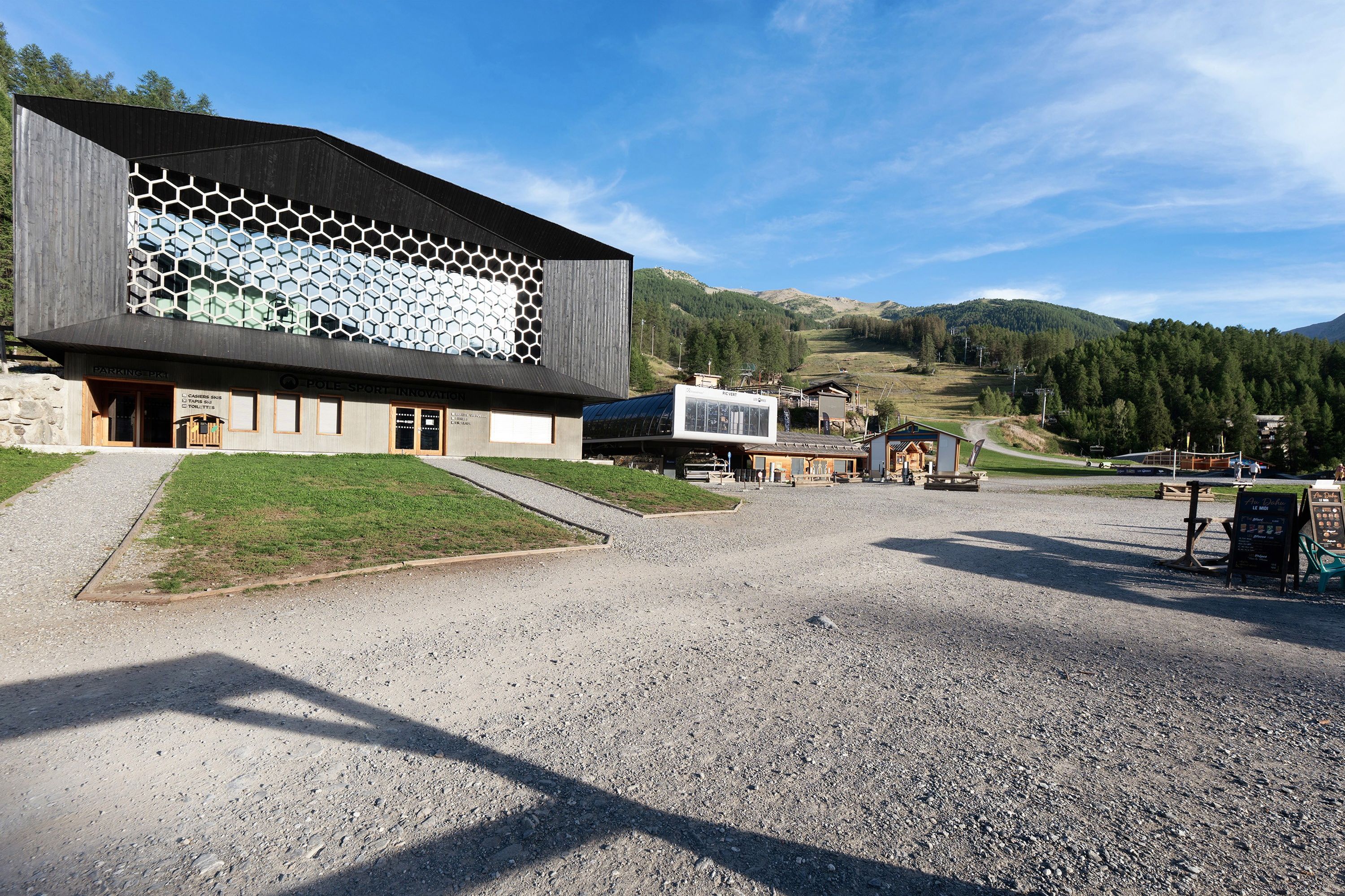 Un bâtiment moderne à la façade hexagonale, avec une allée gravillonnée et un paysage montagneux en arrière-plan.