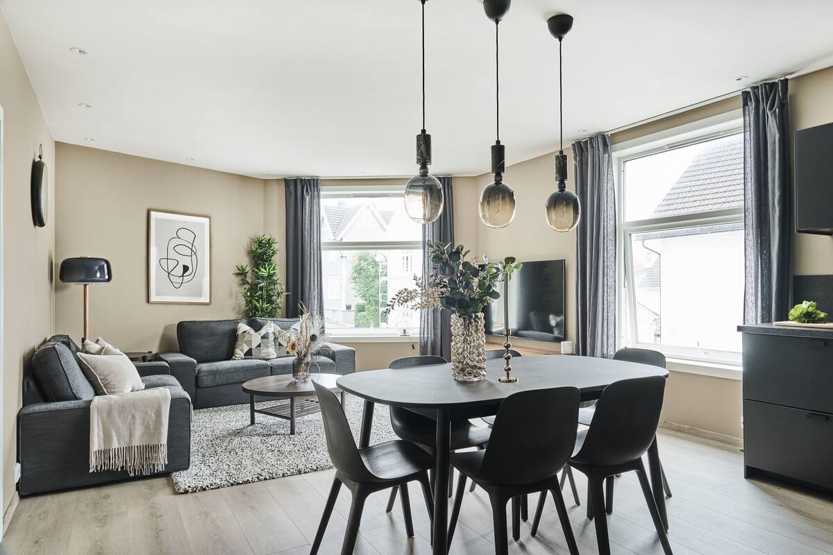 Een moderne woonkamer met een donkere bank, een salontafel en een eethoek met een ronde tafel en stoelen. Er is een groot raam met gordijnen, een plant en een flatscreen televisie die aan de muur is bevestigd.