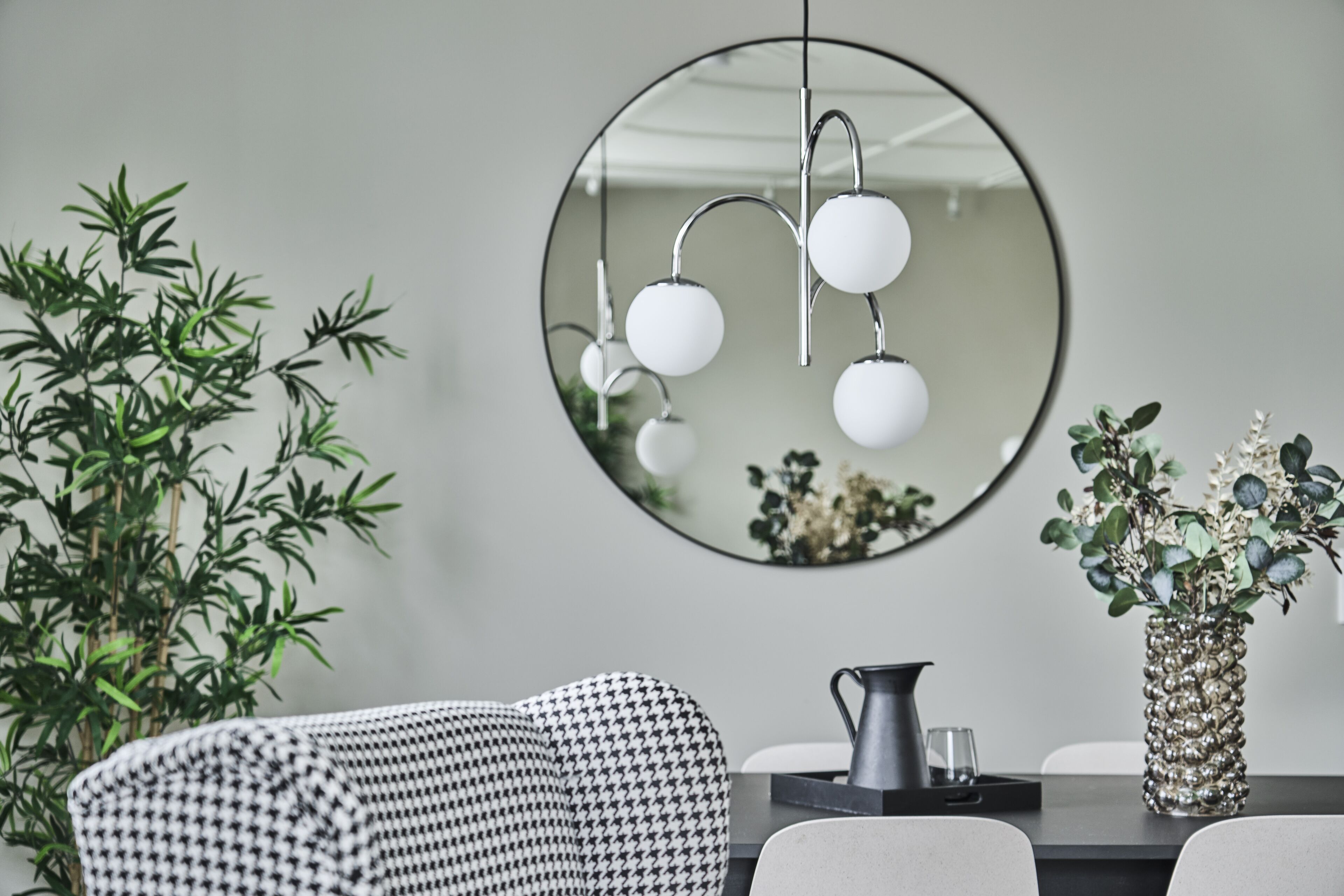 A modern dining area with a round mirror, a houndstooth-patterned sofa, a table with a pitcher, and a decorative vase.
