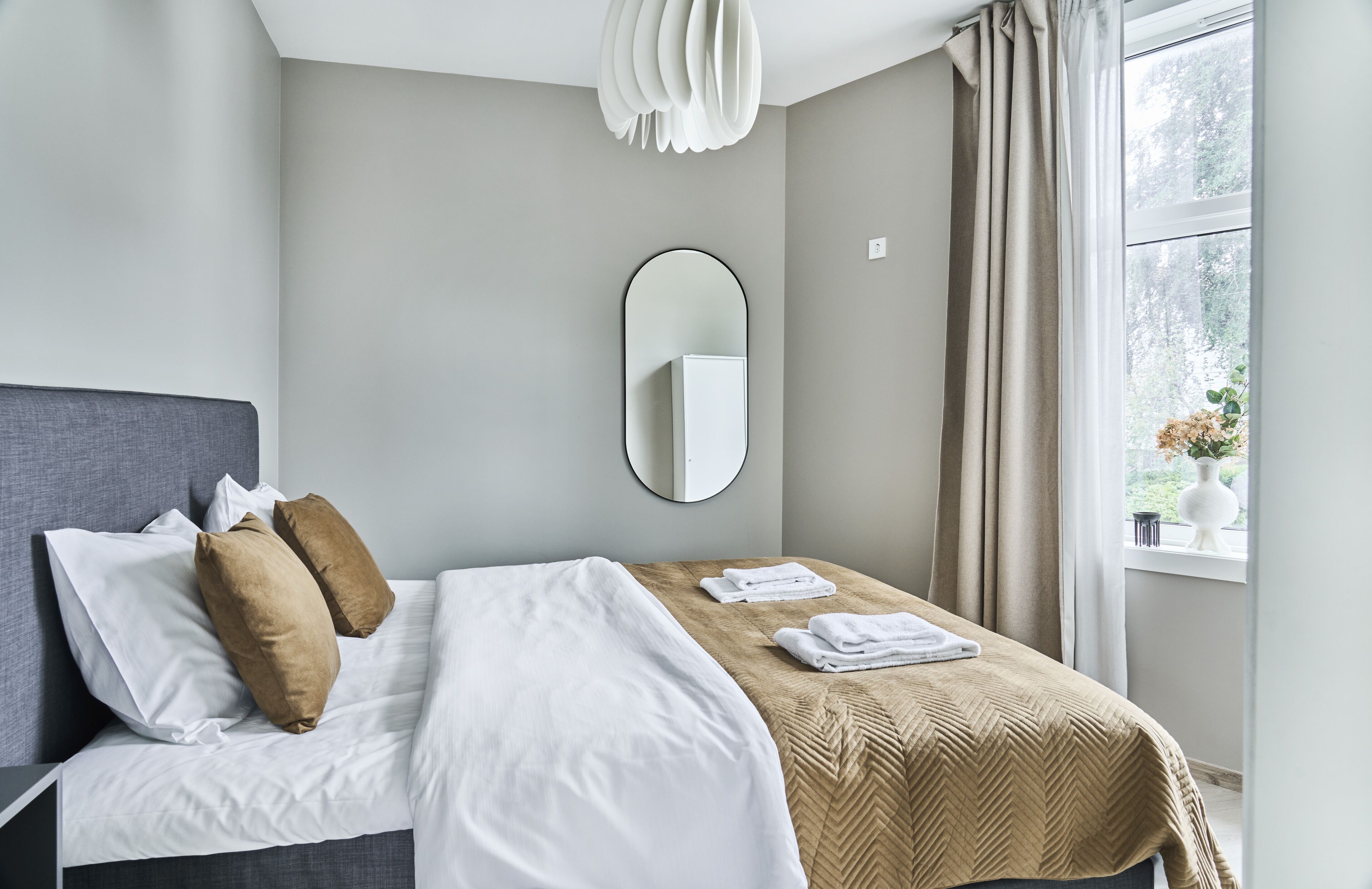 Ein ordentlich bezogenes Bett mit weißen und braunen Kissen, einer beige farbenen Tagesdecke und einem gefalteten Handtuch an der Seite. Ein runder Spiegel hängt an der Wand über dem Bett.