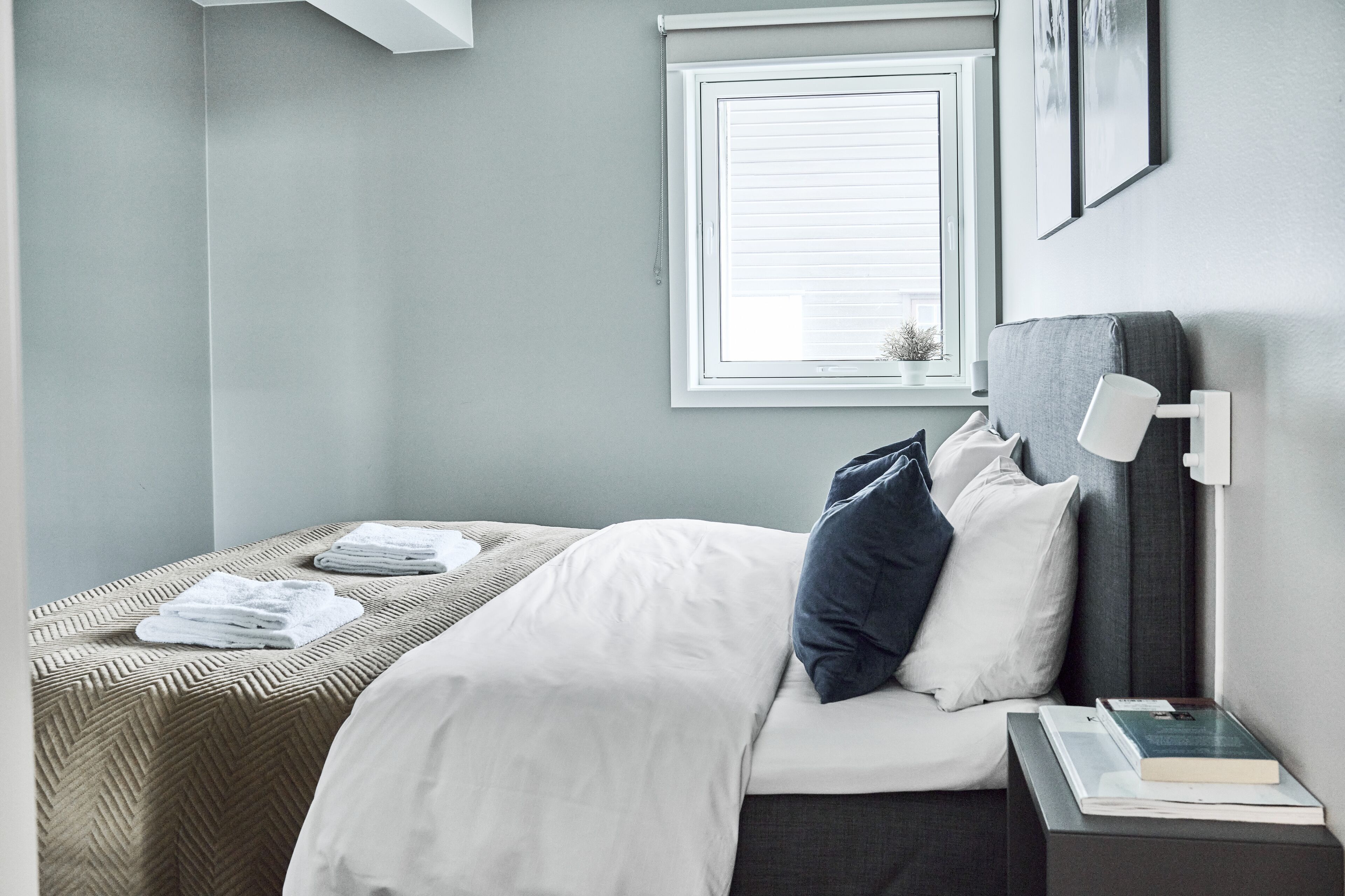 Un lit bien fait, avec du linge de lit blanc et une tête de lit grise, une table de chevet avec un livre et une lampe, et une fenêtre avec des stores.