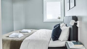 Un lit bien fait, avec du linge de lit blanc et une tête de lit grise, une table de chevet avec un livre et une lampe, et une fenêtre avec des stores.