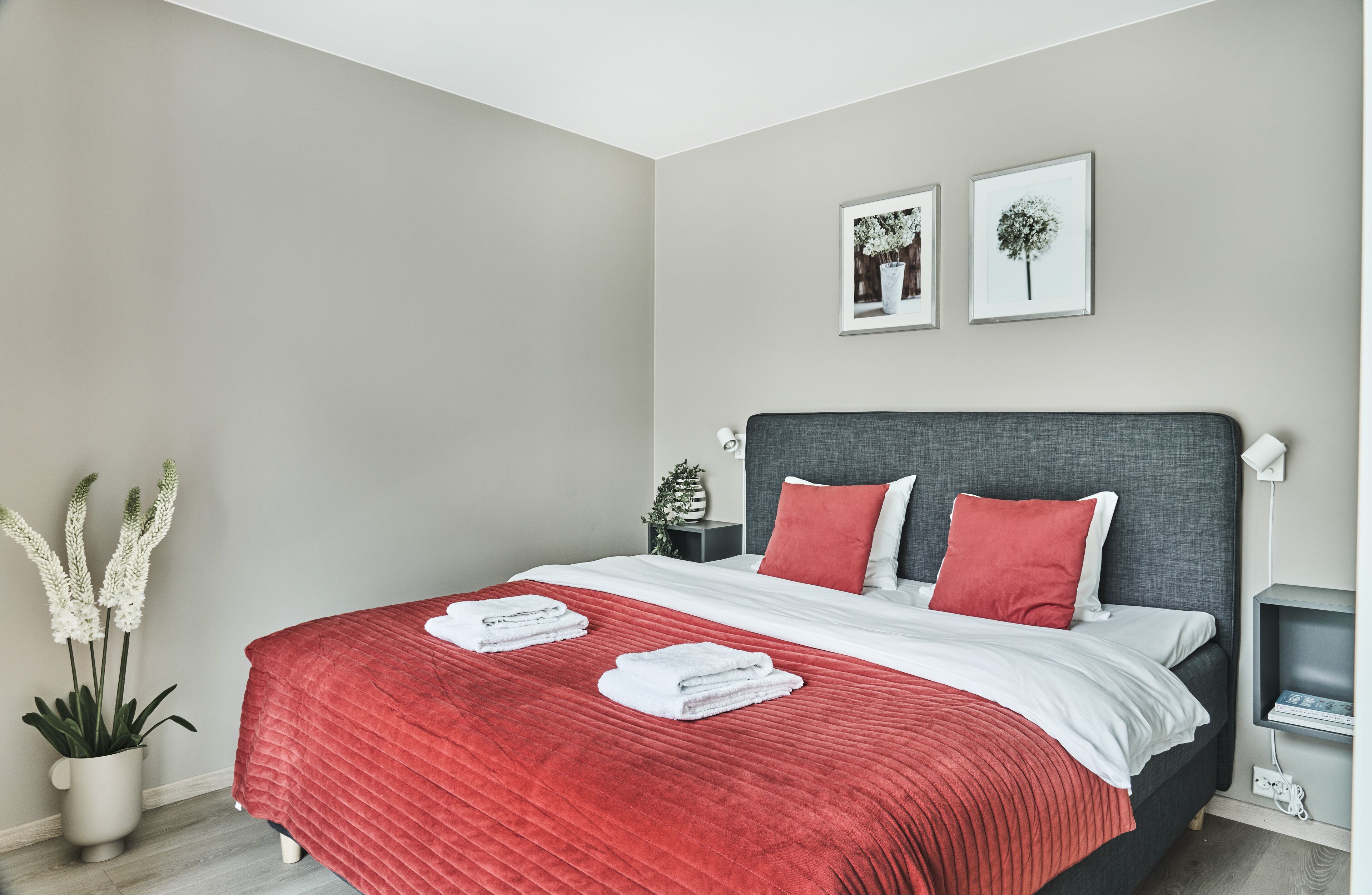 Un dormitorio moderno con una cama grande, ropa de cama roja y blanca, un cabecero gris y dos cuadros en la pared.