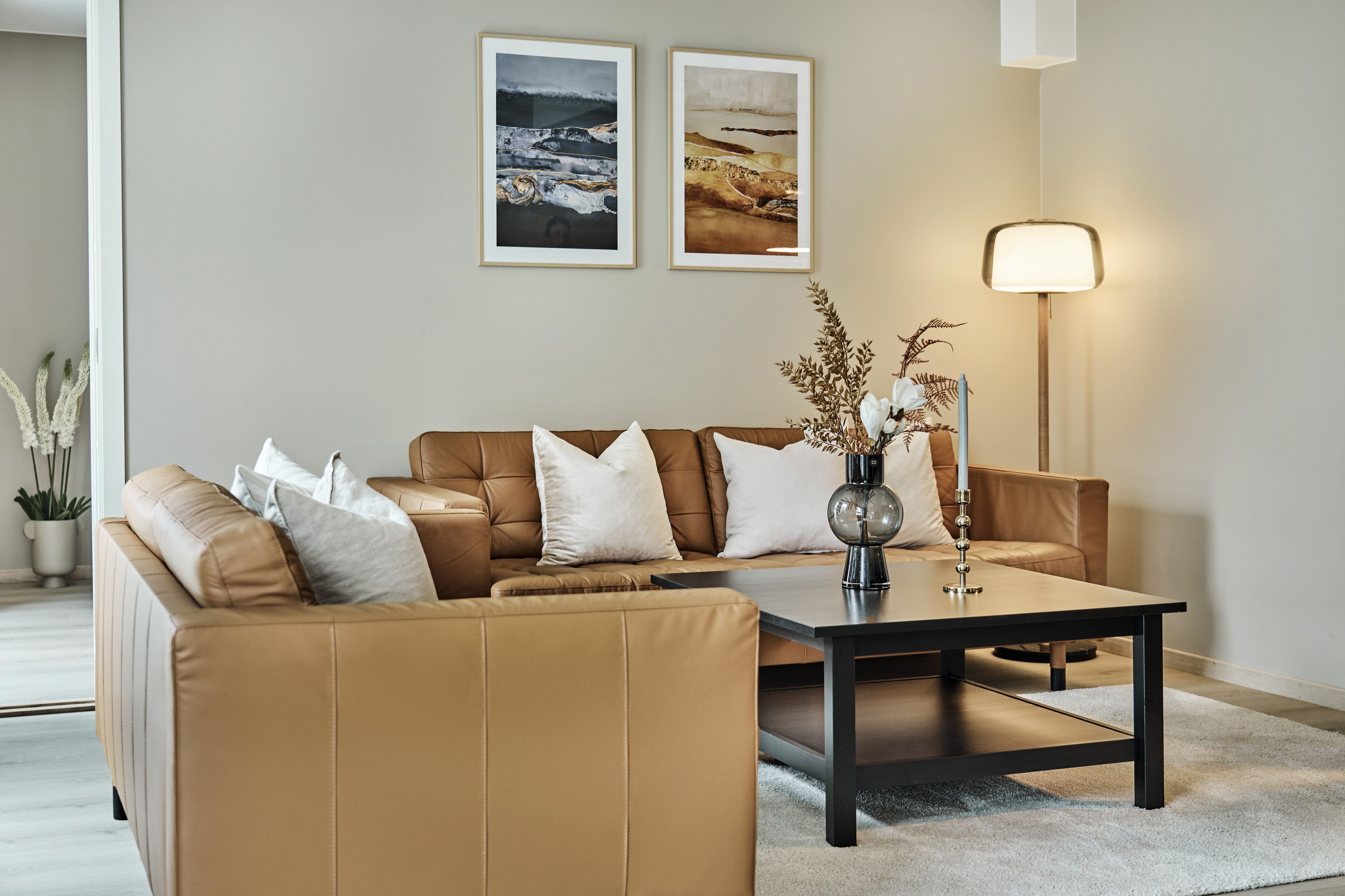 A modern living room with a brown leather sofa, a coffee table, and two framed landscape paintings on the wall.