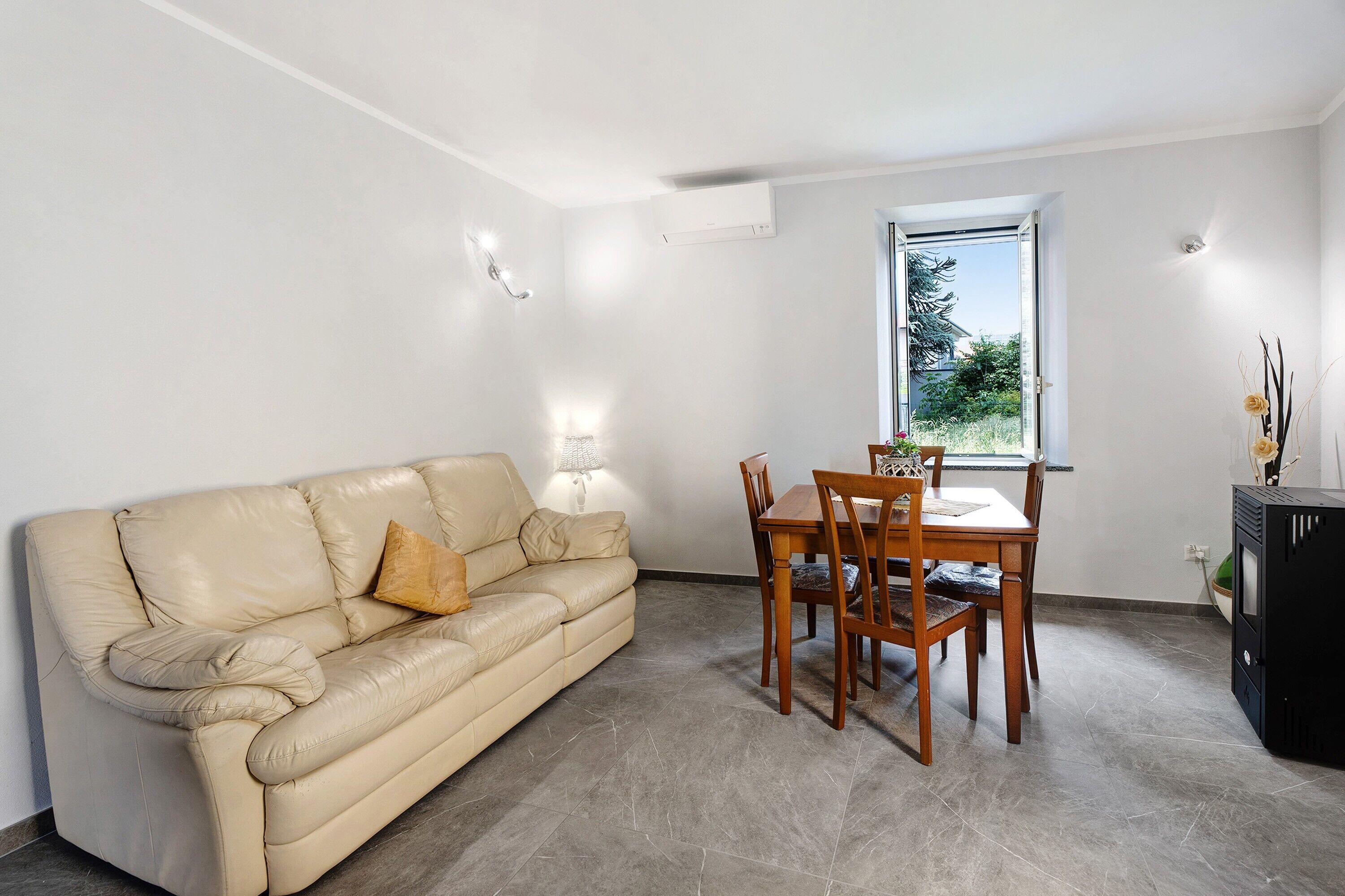 Une pièce avec un canapé en cuir beige, une table à manger en bois avec des chaises, un petit meuble et une fenêtre donnant sur un paysage verdoyant.
