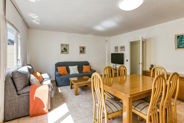 A living room with a sofa, a dining table with chairs, a TV, and framed pictures on the wall.
