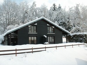 Eine dunkle Holzhütte mit steilem Dach, umgeben von schneebedeckten Bäumen.