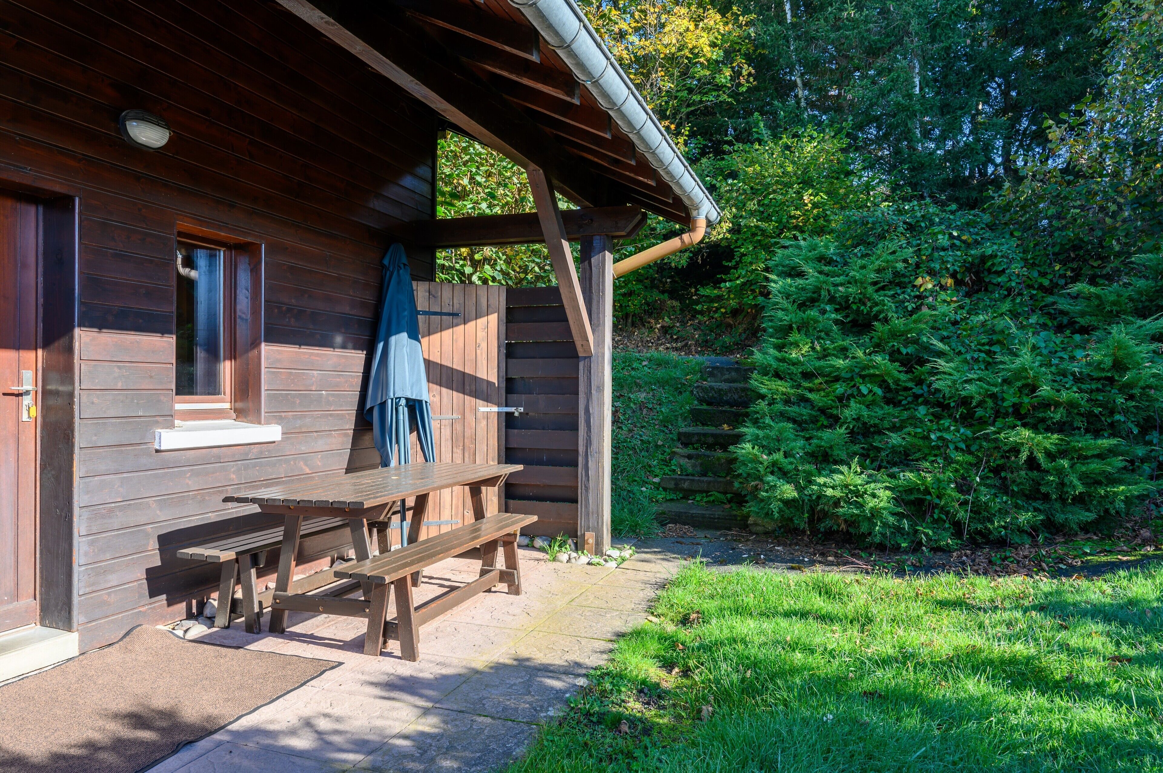 Une cabane en bois avec une table de pique-nique et un espace banc abrité, le tout entouré d’une végétation luxuriante.
