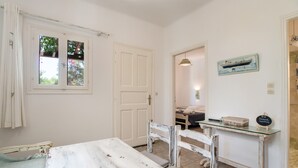 A room with a white table, a bench, a glass-top desk, a door, and a window with curtains. - Holiday Home 'Green Retreats 1 Paros' with Mountain View, Wi-Fi and Air Conditioning (Glifades)