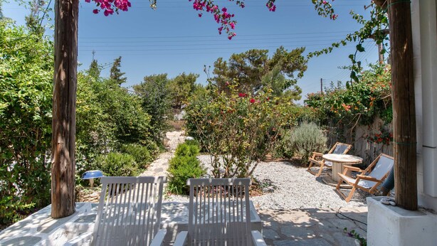 A garden patio with wooden chairs, a white bench, and a pergola with pink flowers. - Holiday Home 'Green Retreats 1 Paros' with Mountain View, Wi-Fi and Air Conditioning (Glifades)