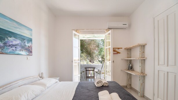 A bedroom with a bed, a painting, a view through an open door, and a shelf with towels. - Holiday Home 'Green Retreats 5 Paros' with Mountain View, Wi-Fi and Air Conditioning (Glifades)