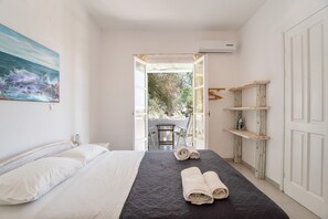A bedroom with a bed, a painting, a view through an open door, and a shelf with towels. - Holiday Home 'Green Retreats 5 Paros' with Mountain View, Wi-Fi and Air Conditioning (Glifades)