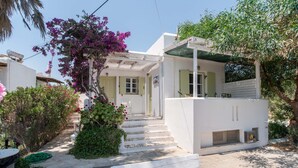 A white building with a covered porch, steps leading to a door, and a garden with purple flowers. - Holiday Home 'Green Retreats 5 Paros' with Mountain View, Wi-Fi and Air Conditioning (Glifades)